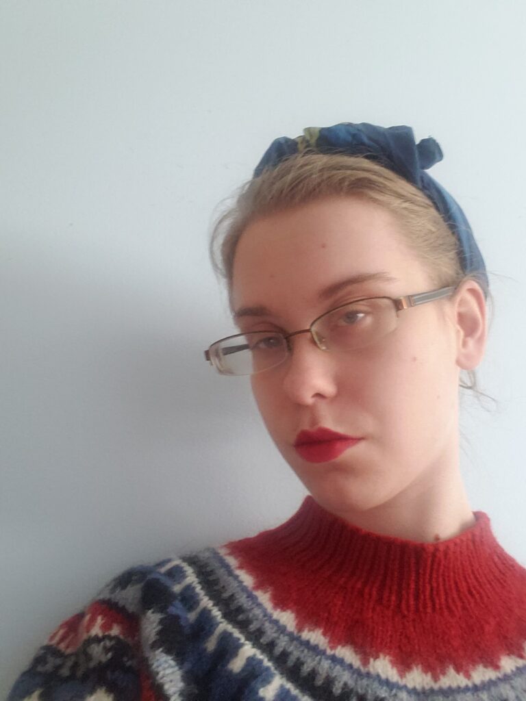 A photo of Grace, wearing a red and black sweater, red lipstick and glasses.