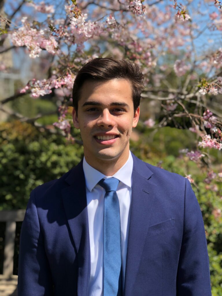 A photo of Kai smiling at the camera and wearing a navy suit. He is standing in front of cherry blossom tree.