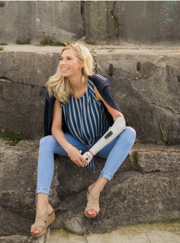 Photo of Nicole, a White woman with blond hair and a white robotic left arm, who is sitting on rocks smiling and wearing jeans and navy and whit striped shirt. 