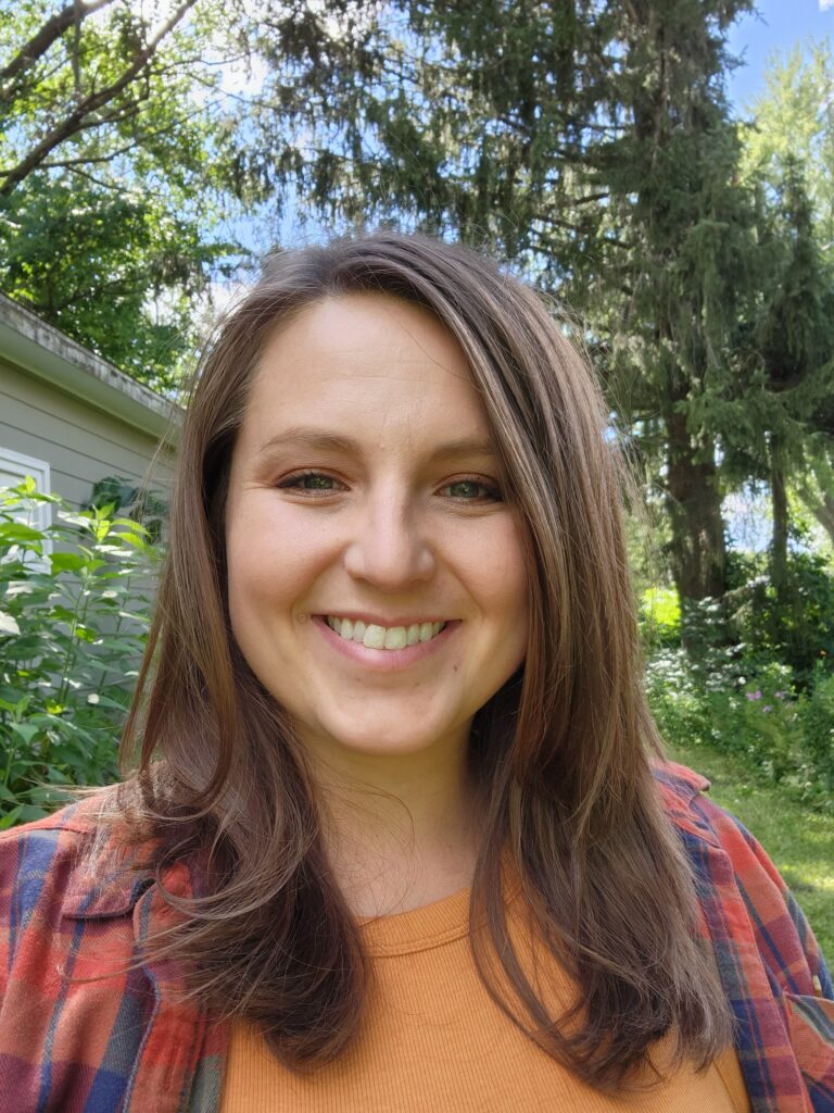 Photo of Allie sitting outside in an orange shirt and flannel. She is smiling at the camera with her mid-length brown hair down. 